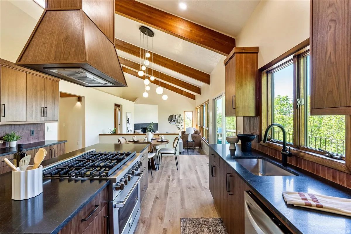 Vaulted kitchen featuring exposed wood beams, wood-finish cabinetry, a large kitchen island with built-in range, dark stone countertops, and a full-height window over the sink