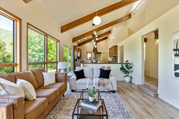 Light-filled living area featuring vaulted ceilings with exposed wood beams, expansive windows, wood-finish flooring, and a contemporary chandelier