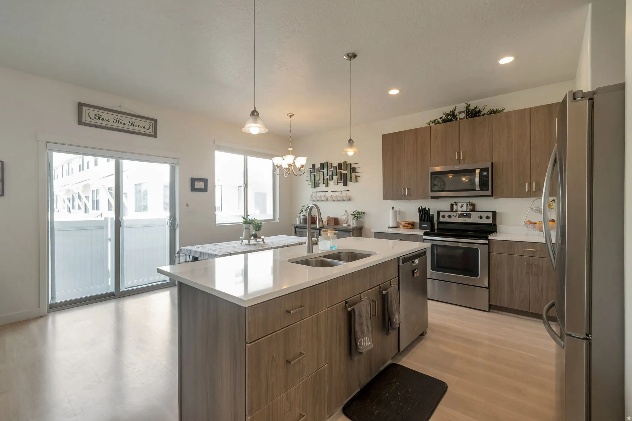 Modern kitchen featuring wood-finish flooring, a central island with dual sinks and pendant lighting, stainless steel appliances, light-toned countertops, and wood-finish cabinetry