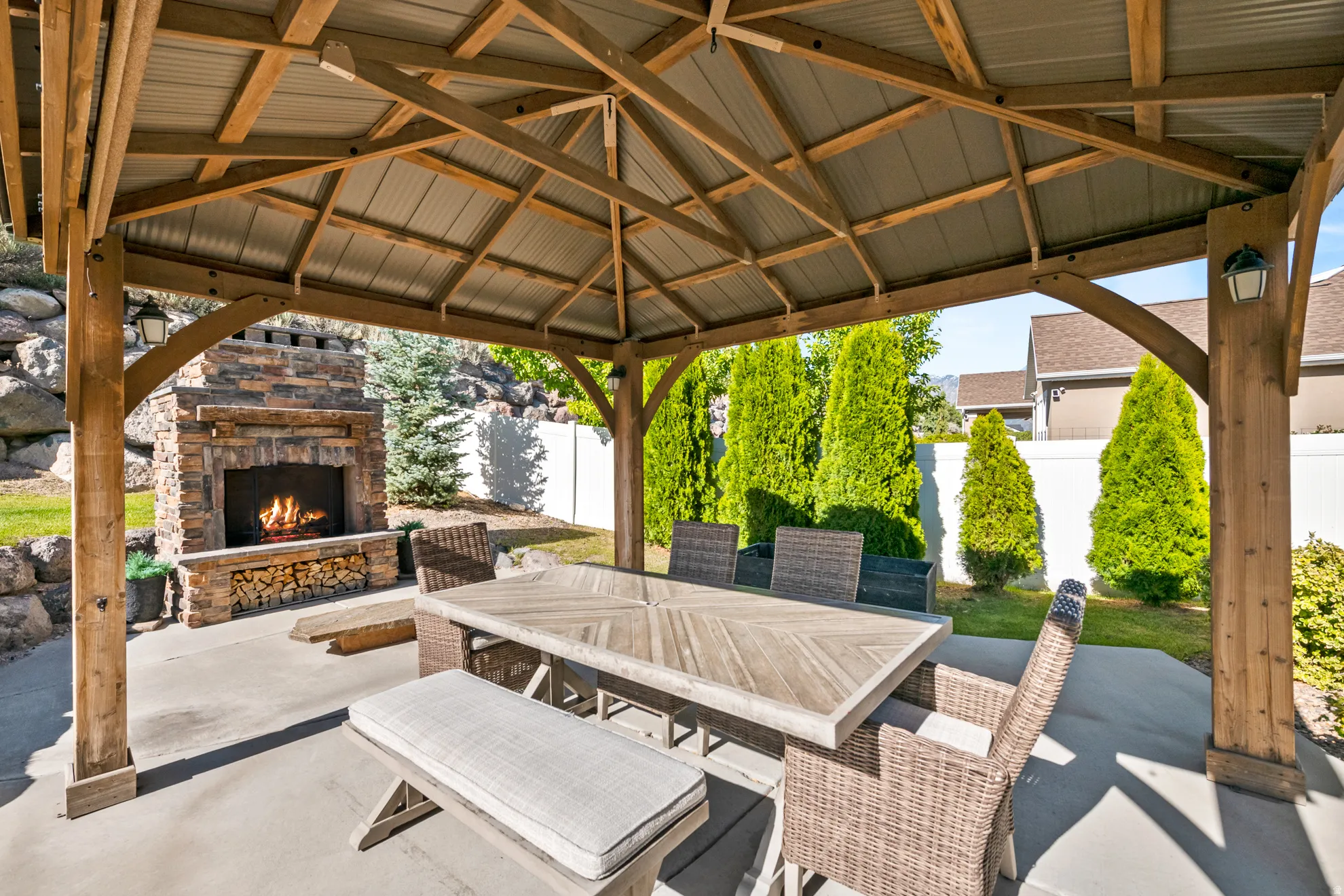 Covered patio featuring a wood-beam gazebo, stone fireplace, concrete flooring, and surrounding privacy landscaping