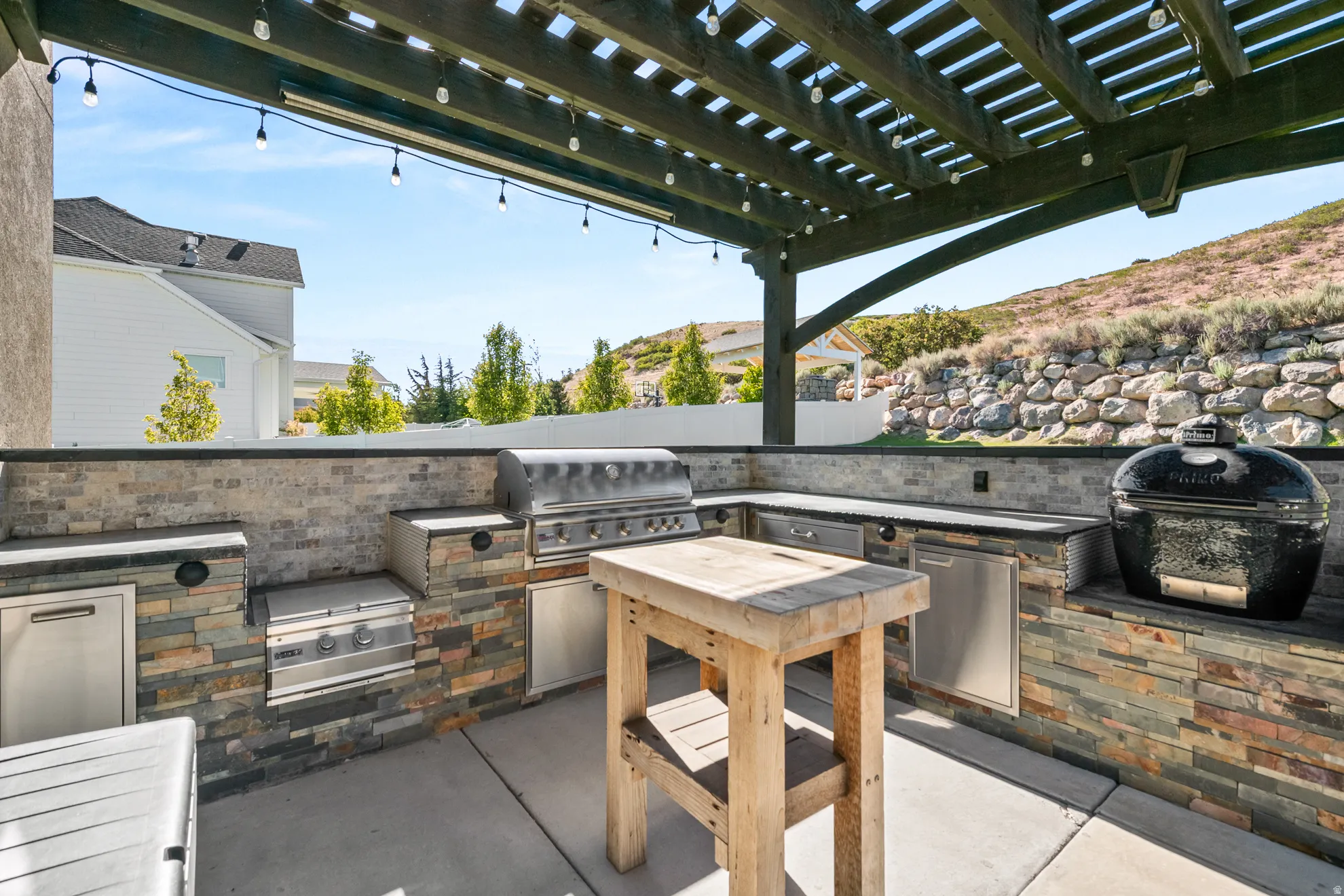 Outdoor kitchen featuring a dark wood pergola, string lighting, stacked stone facade, stainless steel appliances, and a concrete patio