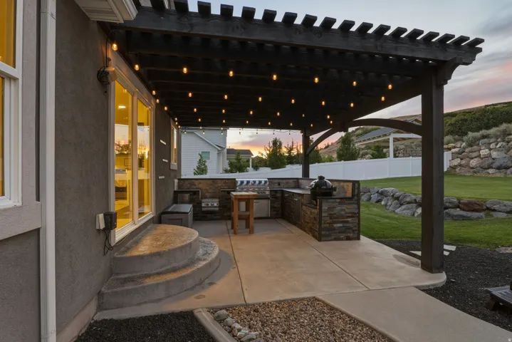 Expansive outdoor living area featuring a dark wood pergola, built-in outdoor kitchen with stone veneer, concrete patio, and a manicured lawn