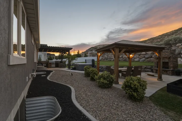 Expansive backyard featuring a wood-frame gazebo, concrete patio, built-in stone fireplace, and a stone retaining wall