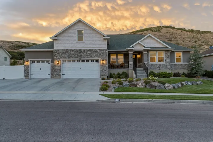 Stone and shingle exterior with a covered porch, established landscaping, two-car garage, and a concrete driveway