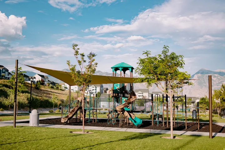 Community playground featuring multiple slides, climbing structures, a shade canopy, and rubberized ground cover, all set within a landscaped green space with mountain scenery
