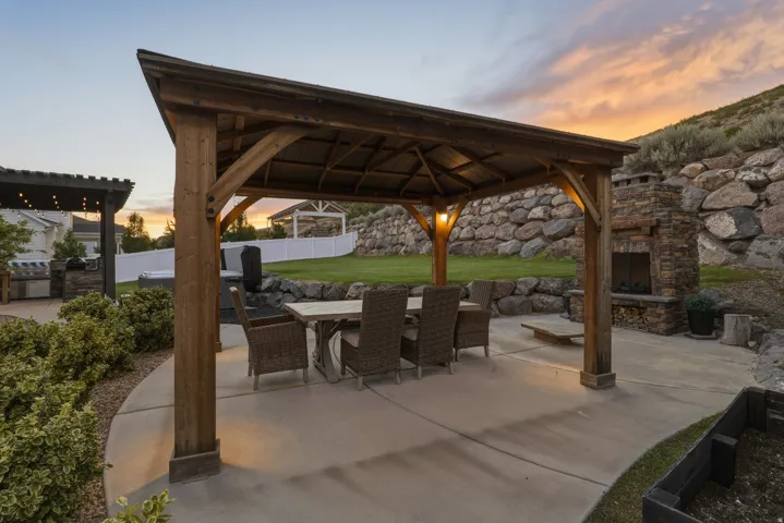 Wooden gazebo with built-in lighting, stone-clad outdoor fireplace, concrete patio, and a rock retaining wall