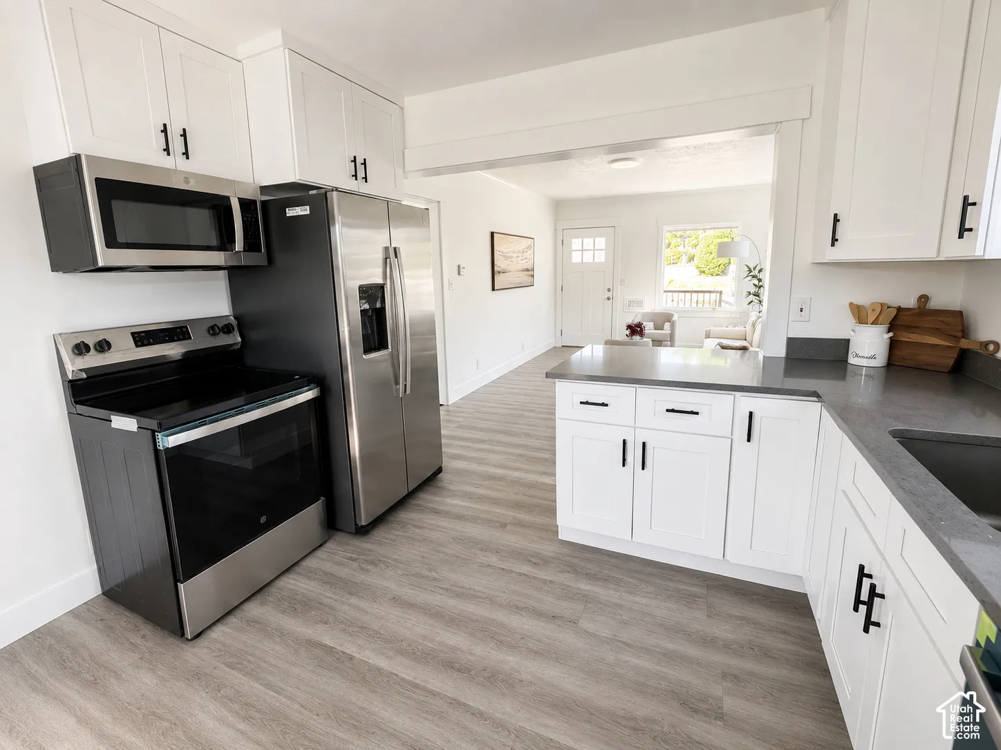 Modern kitchen featuring light wood-finish flooring, white shaker-style cabinetry with matte black hardware, stainless steel appliances, and dark countertops