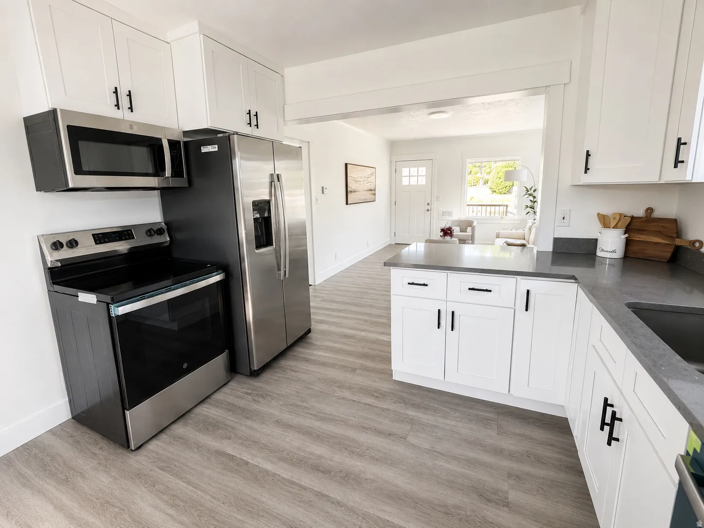 Modern kitchen featuring light wood-finish flooring, white shaker-style cabinetry with matte black hardware, stainless steel appliances, and dark countertops