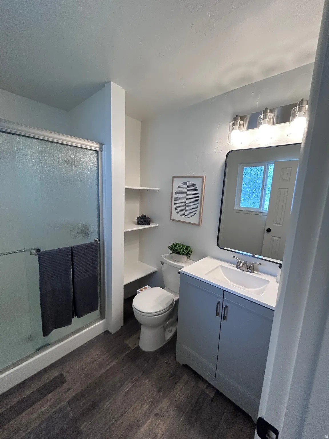 Bathroom featuring a walk-in shower with glass enclosure, a single vanity with grey cabinetry and white countertop, wood-finish flooring, built-in shelving, and a toilet