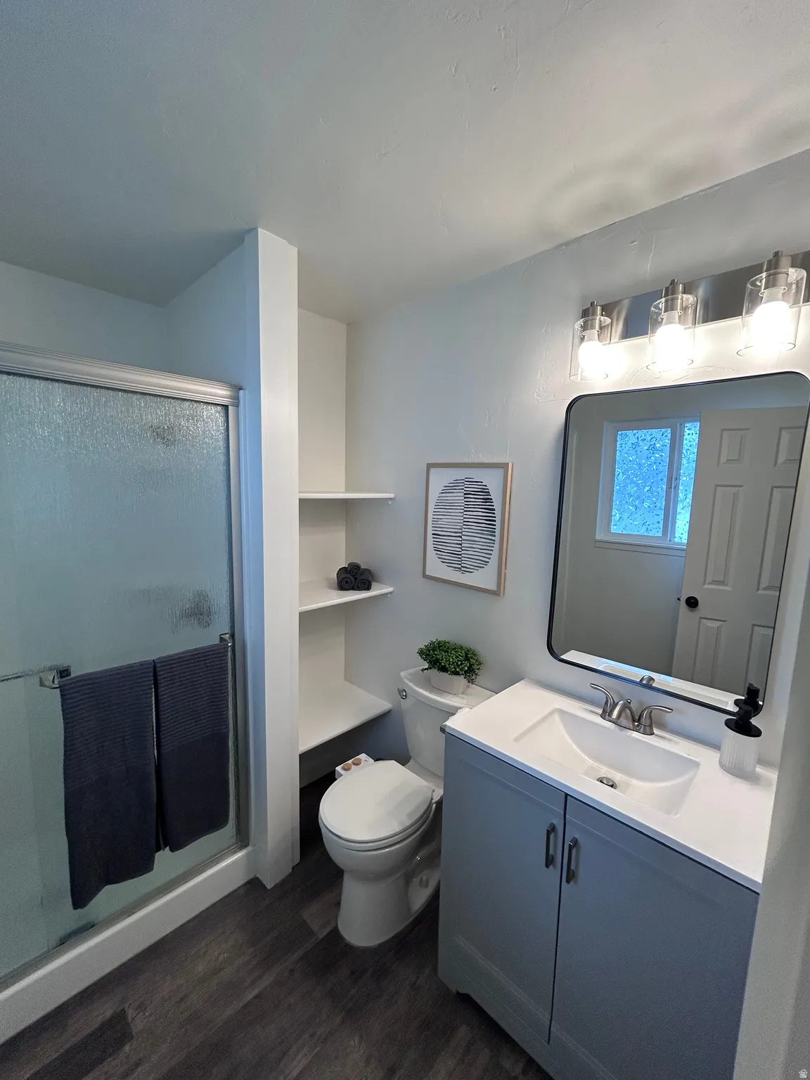 Bathroom featuring a walk-in shower with glass enclosure, white vanity with integrated sink, contemporary vanity lighting, built-in shelving, and wood-finish flooring