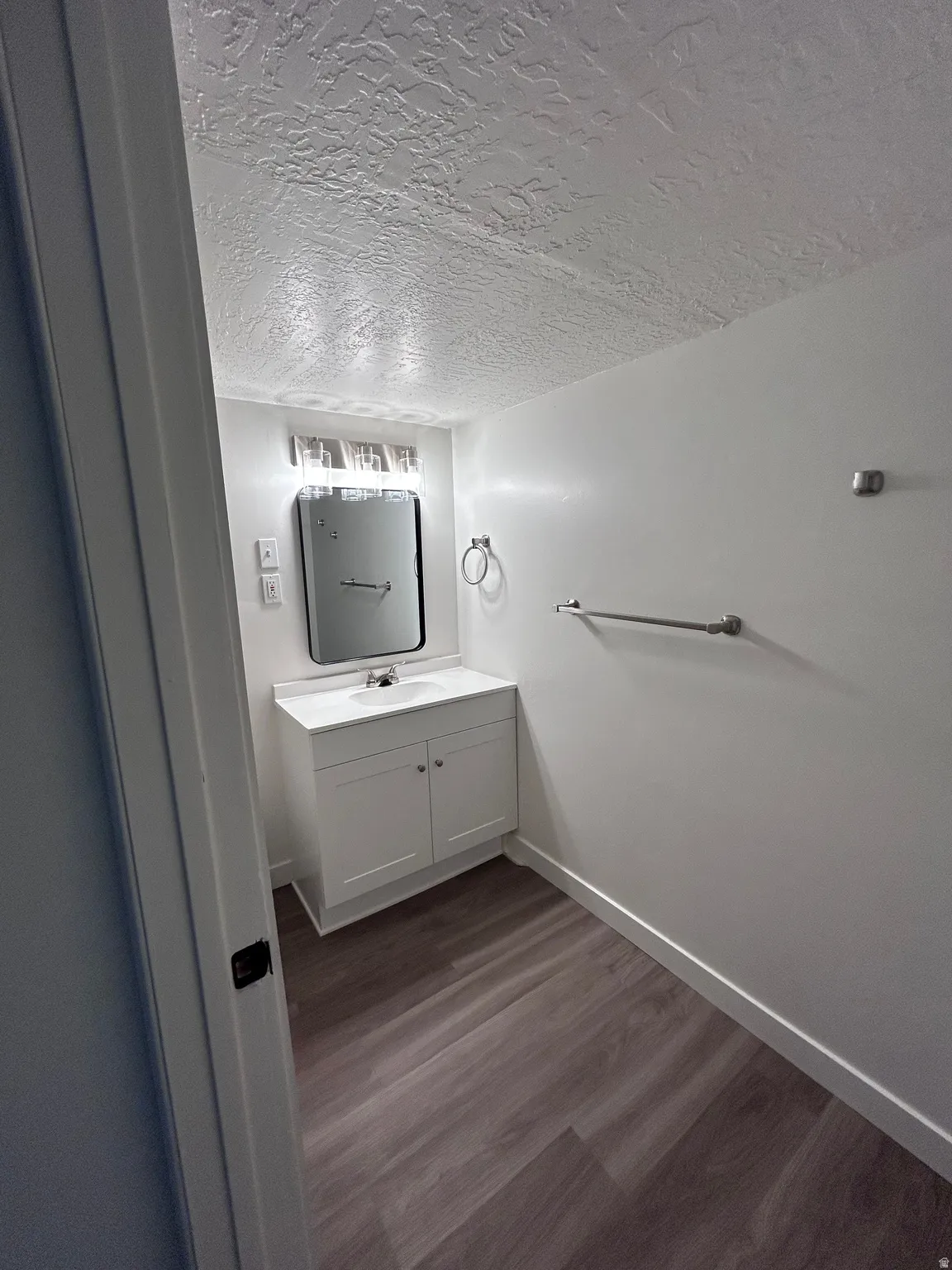 Bathroom featuring a single vanity with a white countertop and cabinet storage, wood-finish flooring, a wall-mounted mirror, and overhead lighting