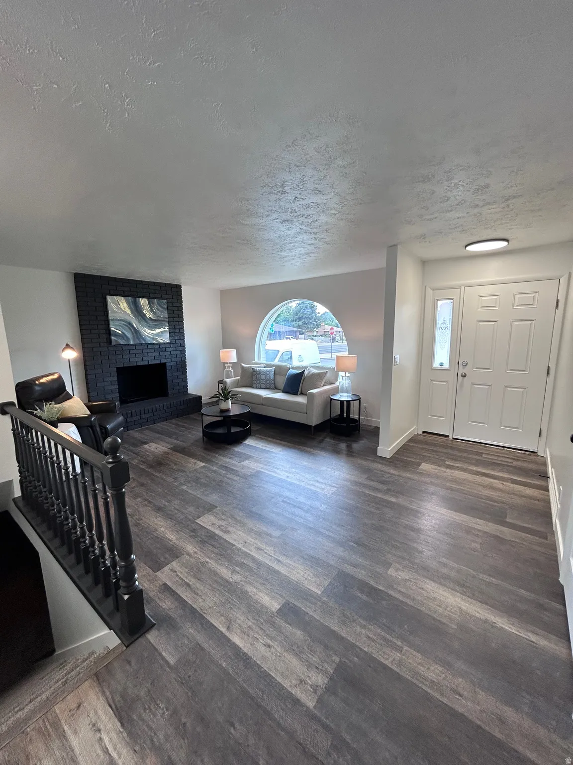 Spacious living area featuring wood-finish flooring, a dark brick fireplace, a large arched window, and a dual-pane entry door with sidelights