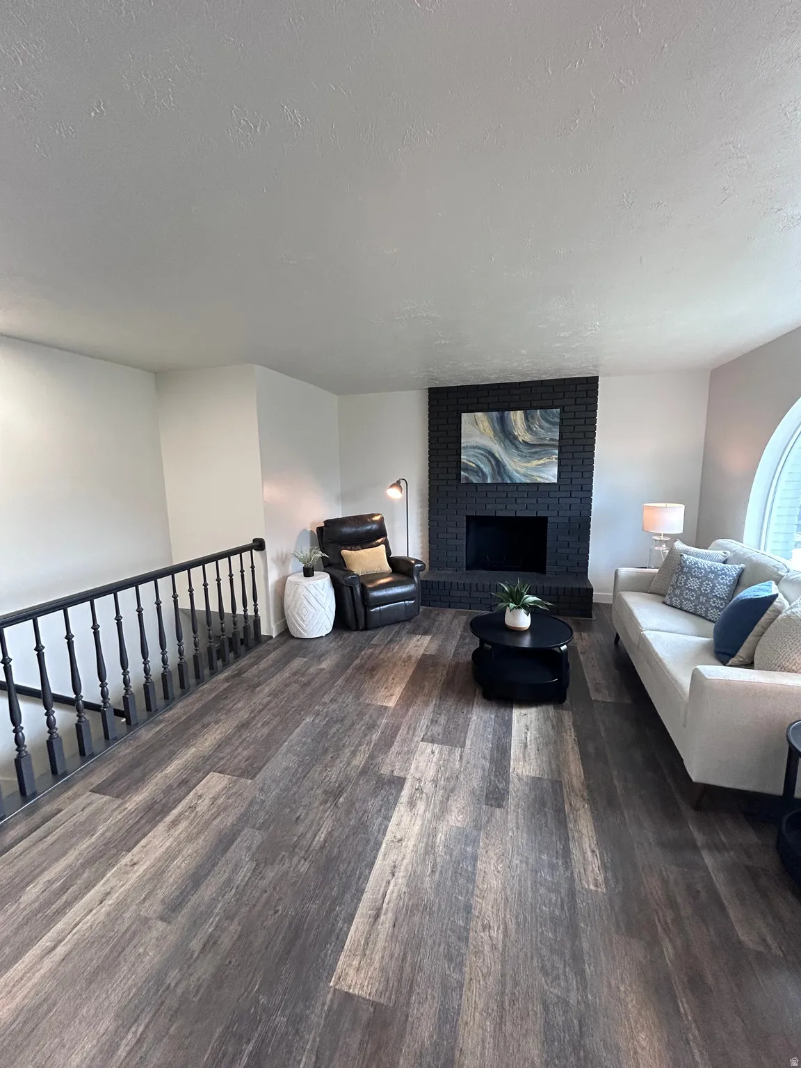 Spacious living area featuring wood-finish flooring, a dark brick fireplace, and a black staircase railing