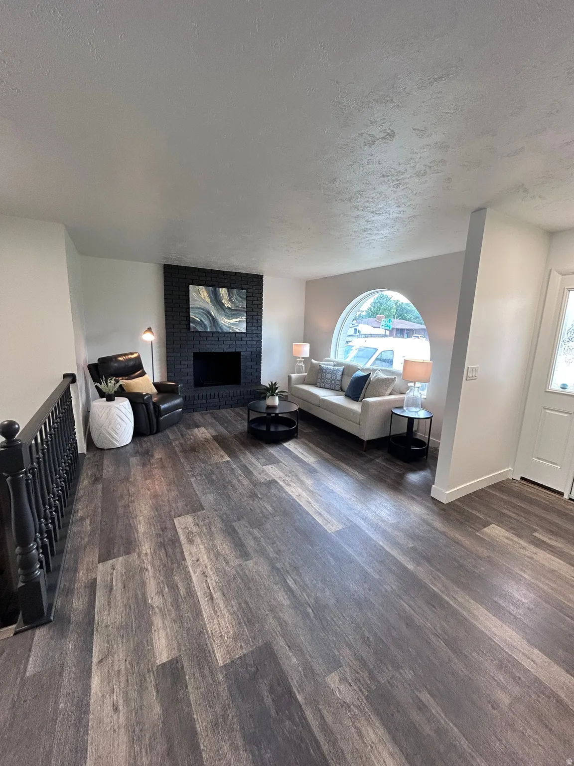 Spacious living area featuring wood-finish flooring, a black brick fireplace, and a large arched window