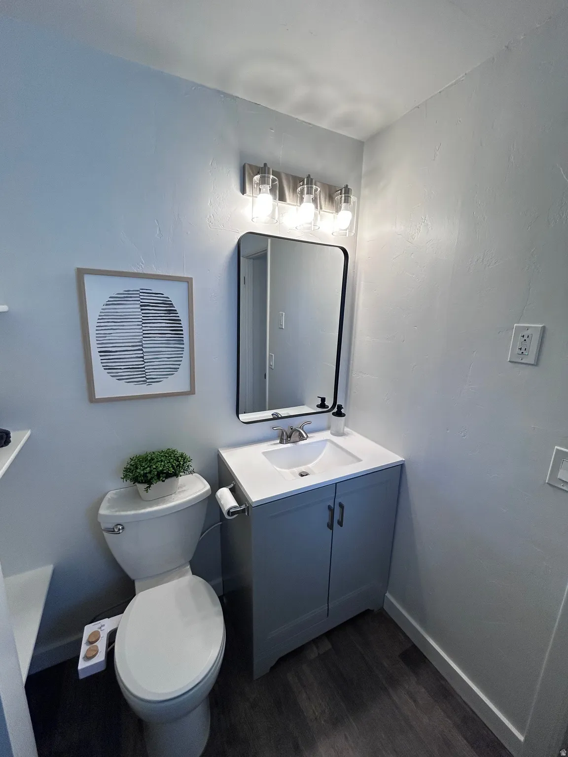 Powder room featuring a single-sink vanity with white countertop and gray cabinetry, dark wood-finish flooring, a contemporary black-framed mirror, and a three-light vanity fixture