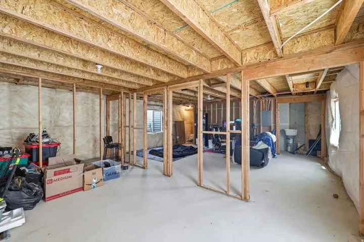 Unfinished basement with exposed ceiling joists and wood framing, featuring concrete flooring, insulated exterior walls, and a visible utility sink