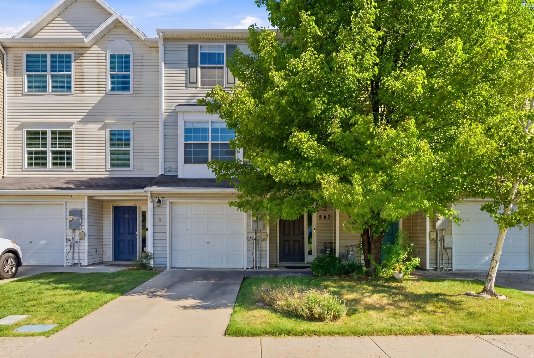 Multi-story townhome featuring light-colored siding, a prominent front entry with a blue door, an integrated garage, and a lush green lawn