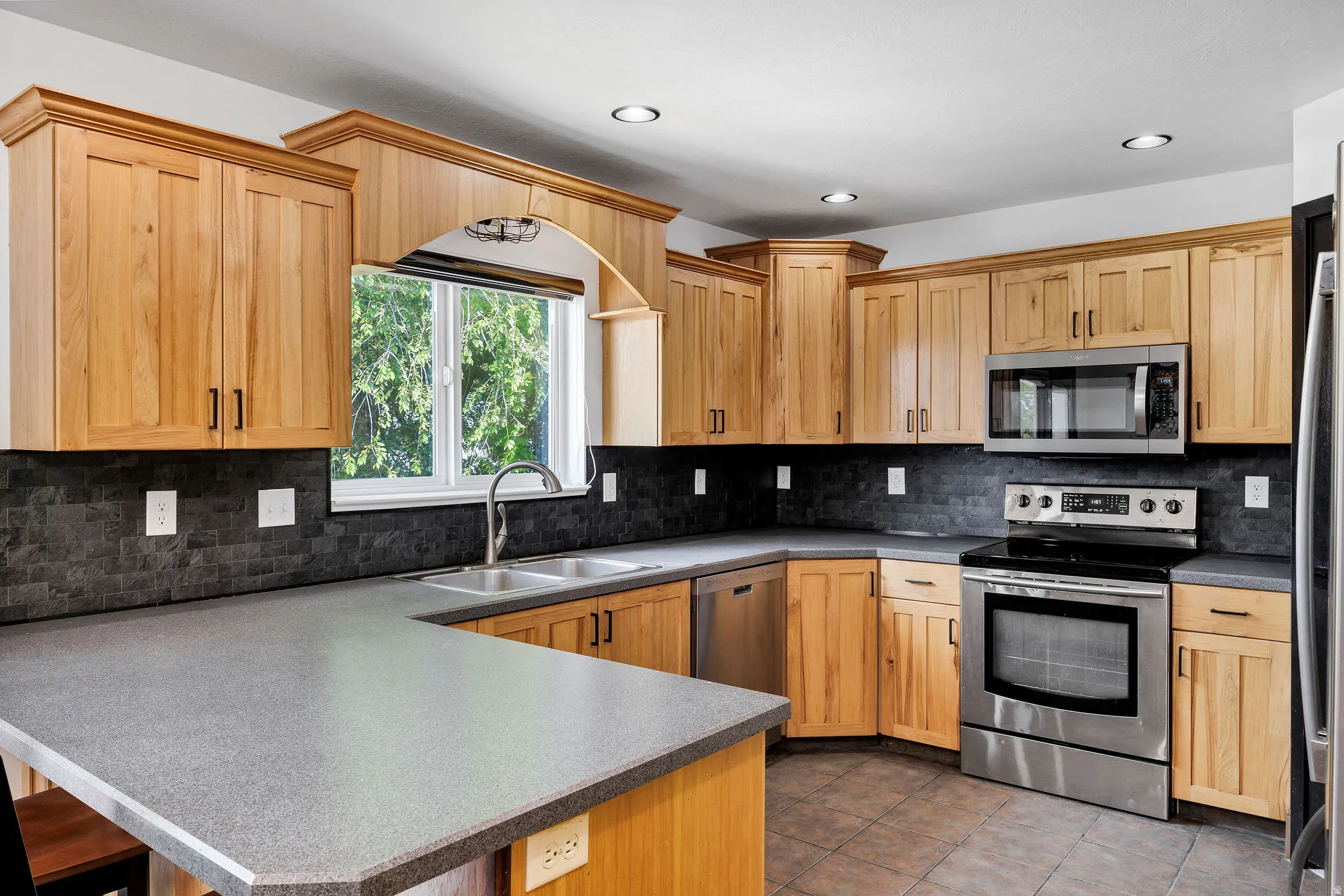 Kitchen featuring light wood cabinetry, stainless steel appliances, a double basin sink, stone-look countertops, and a dark tile backsplash