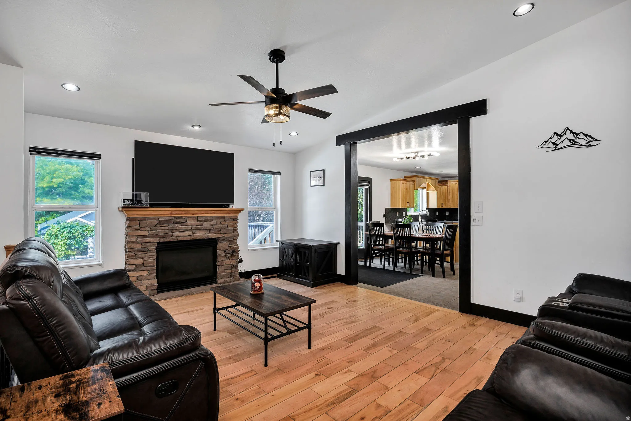 Living area featuring a stone-clad fireplace with wood mantle, light wood-finish flooring, a ceiling fan with integrated lighting, recessed lighting, and an expansive doorway opening to an additional room