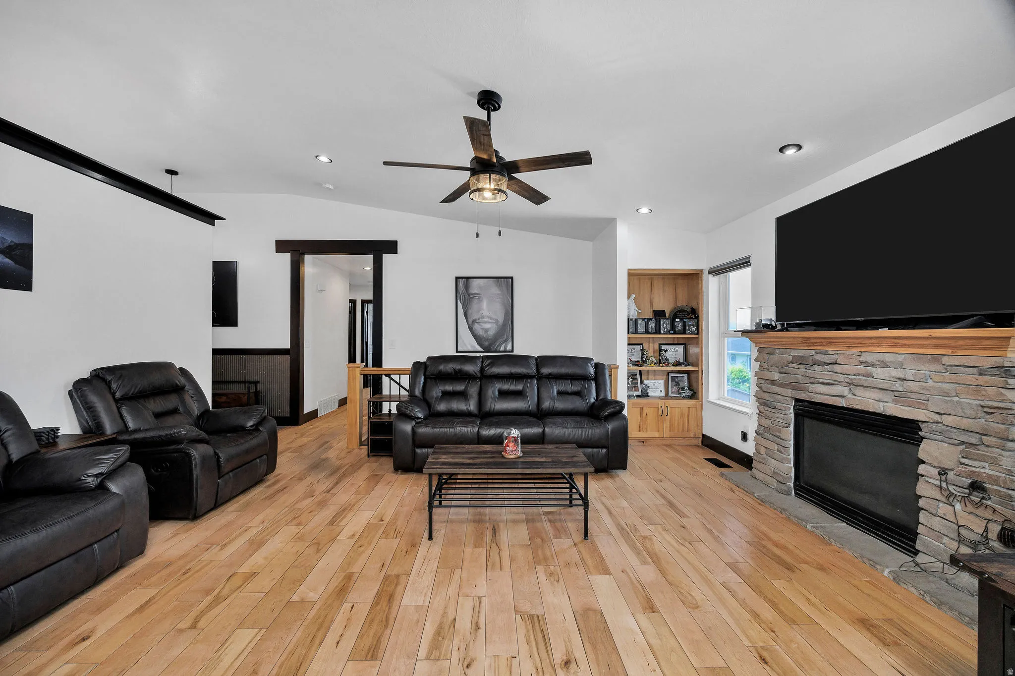 Spacious living area featuring wood-finish flooring, a stone-clad fireplace with a wood mantle, a ceiling fan, recessed lighting, and a built-in wooden shelving unit