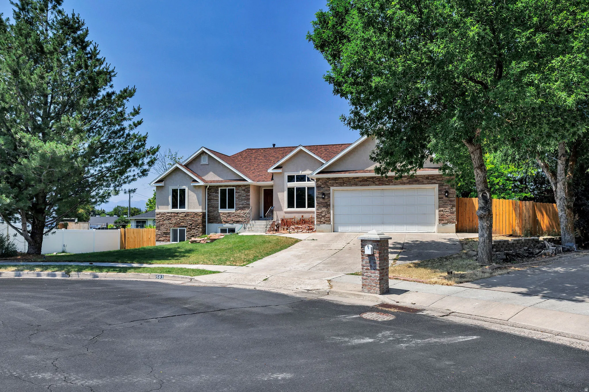 Two-story residence featuring a stone and stucco facade, a two-car garage, mature landscaping, a concrete driveway, and a red shingle roof
