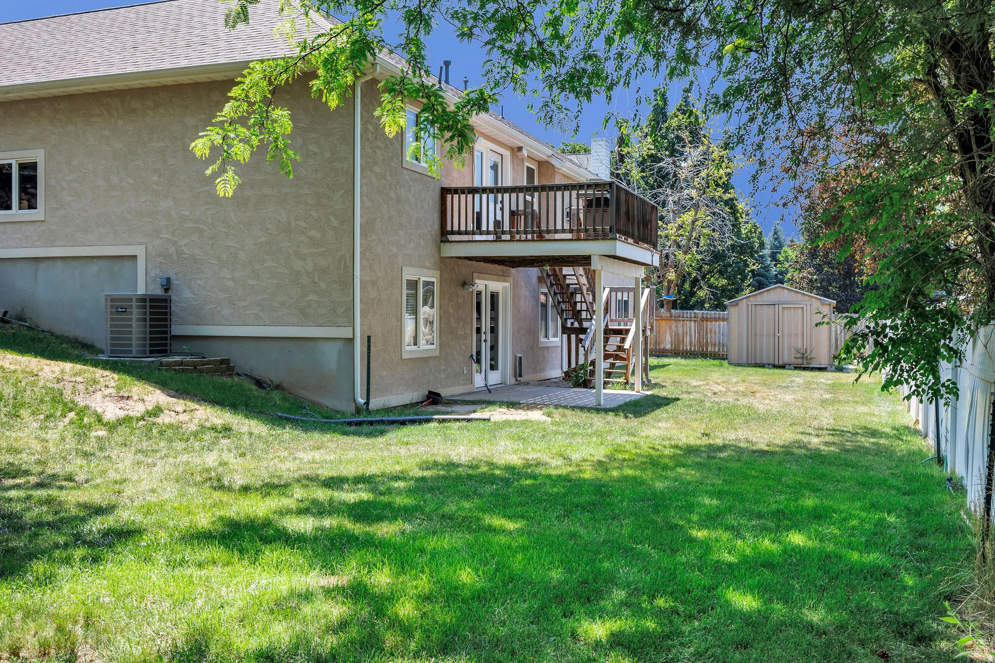 Spacious backyard featuring a two-story residence with stucco siding, an upper-level wood deck, a lower-level patio, and a detached storage shed