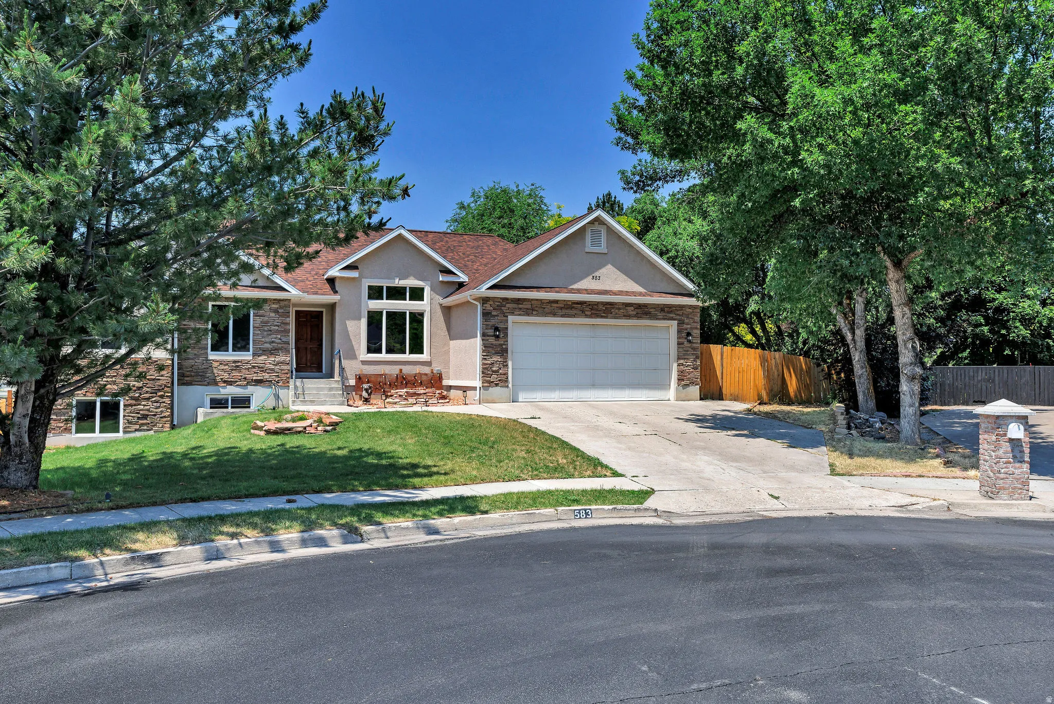 Stucco and stone facade home featuring a two-car garage, mature trees, and a sloped front lawn