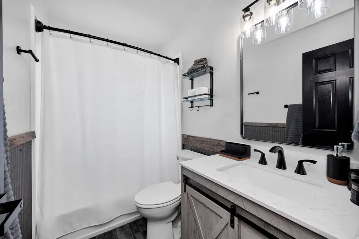 Bathroom featuring a shower-tub combination with a curved rod, a vanity with a barn door style cabinet, a white countertop with an integrated sink, a rectangular framed mirror, and a three-light vanity fixture
