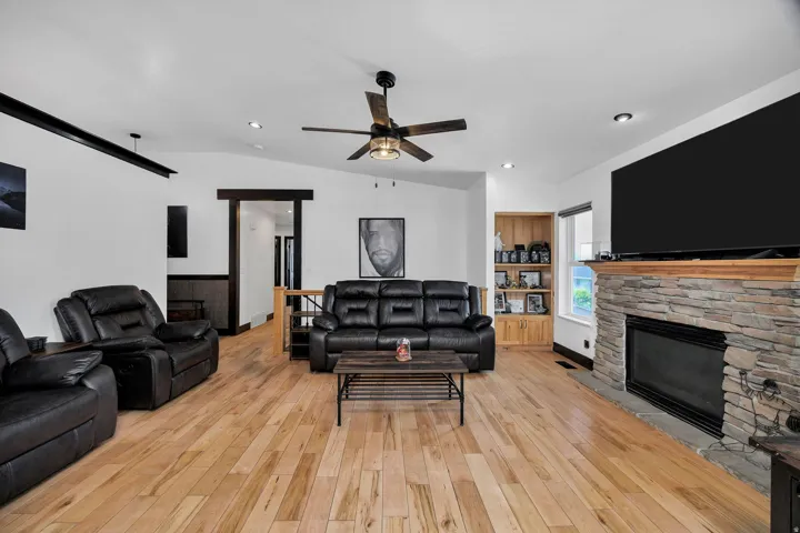 Spacious living area featuring wood-finish flooring, a stone-clad fireplace with a wood mantle, a ceiling fan, recessed lighting, and a built-in wooden shelving unit