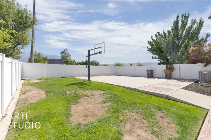 Expansive backyard featuring a concrete pad with a basketball hoop, a manicured lawn, and white privacy fencing