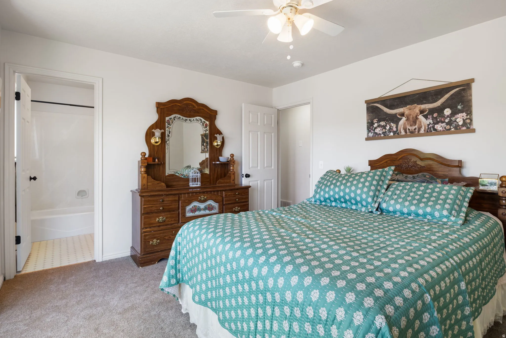 Carpeted bedroom with a built-in ceiling fan, white walls, and a recessed doorway leading to a bathroom featuring a white tub and tiled flooring