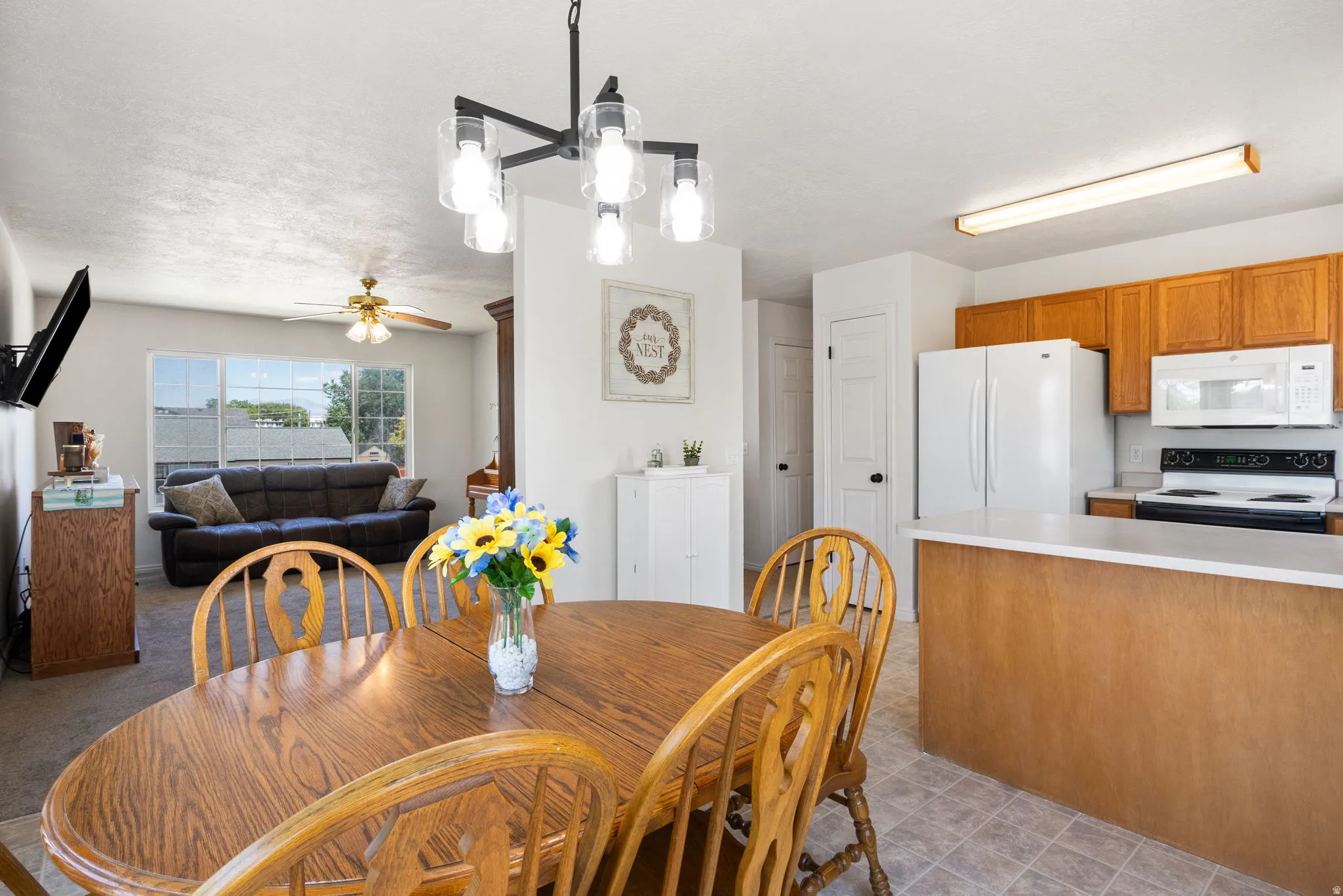 Open-concept living area featuring a dining space with a modern chandelier, a living room with a large window, a kitchen with wood cabinetry and a peninsula, and tile flooring