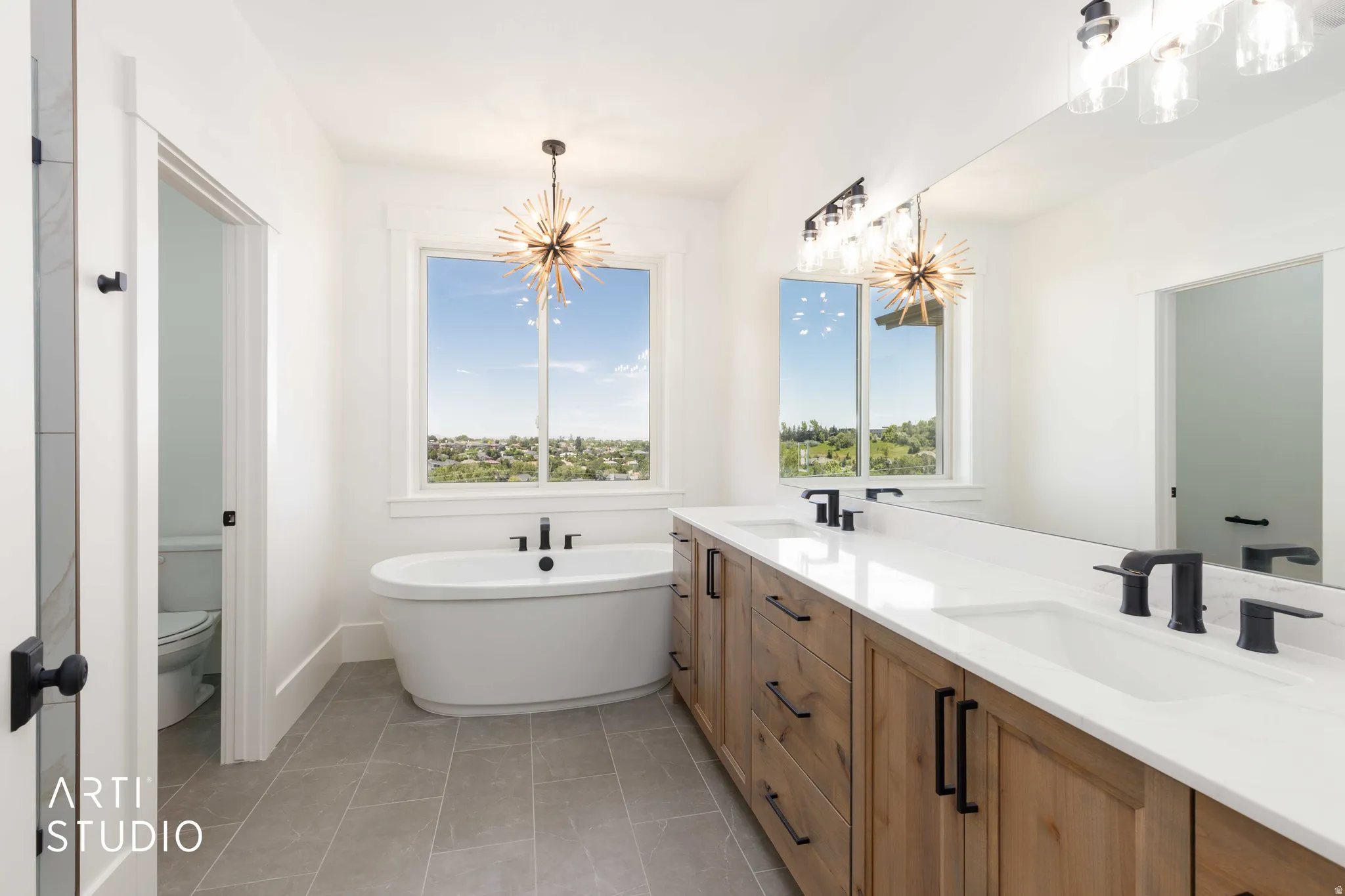 Spacious bathroom featuring a freestanding soaking tub with a floor-mounted matte black faucet, a large dual vanity with wood-finish cabinetry and white countertops, rectangular undermount sinks with matte black faucets, and rectangular floor tiles