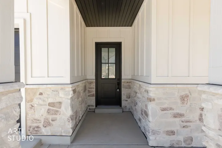 Covered entry featuring a dark-toned door with glass lites, stone veneer accents, vertical board-and-batten siding, and a dark-toned soffit