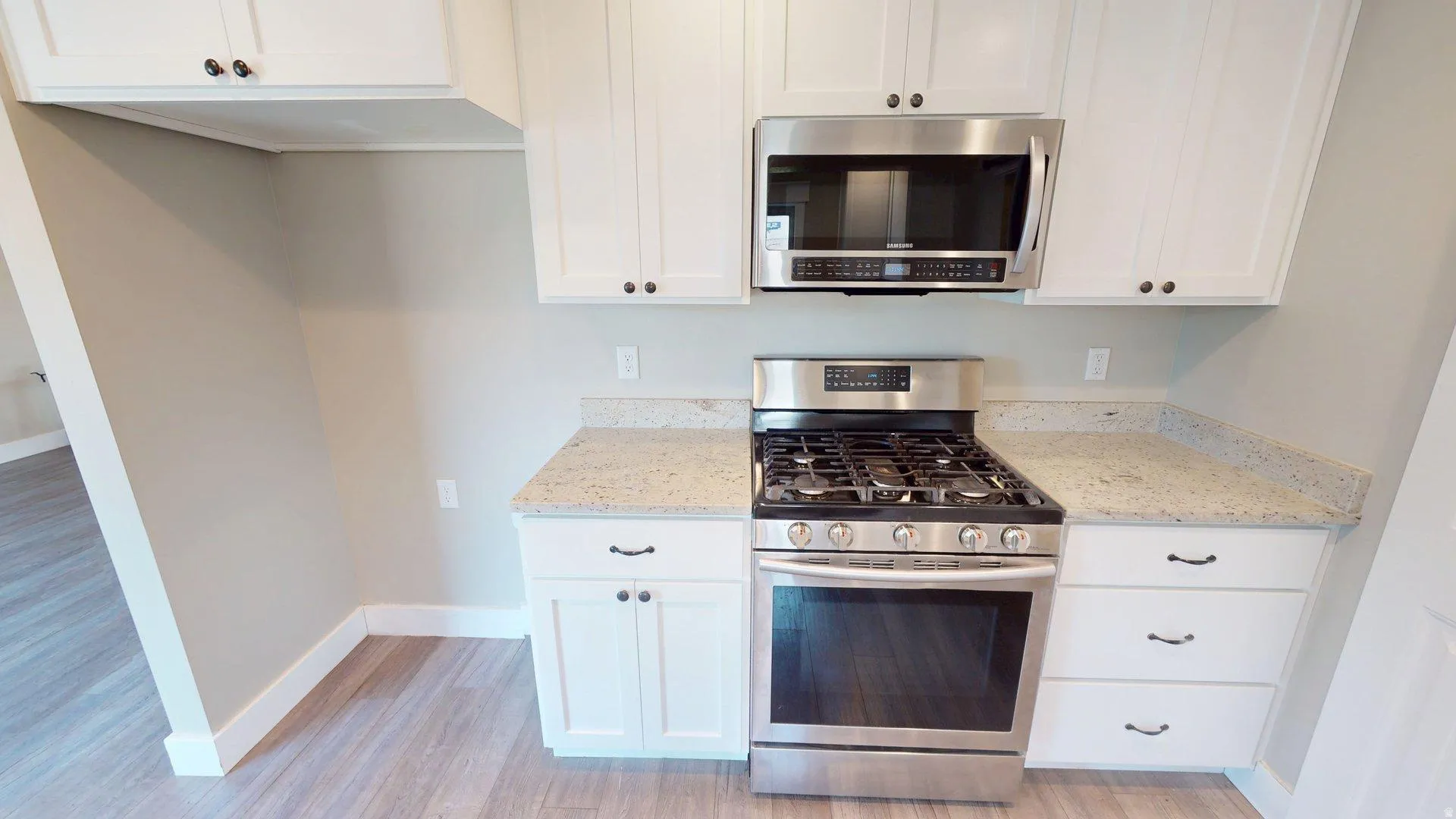 Kitchen featuring stainless steel range with gas cooktop, stainless steel microwave, white shaker-style cabinetry, light-toned countertops, and wood-finish flooring