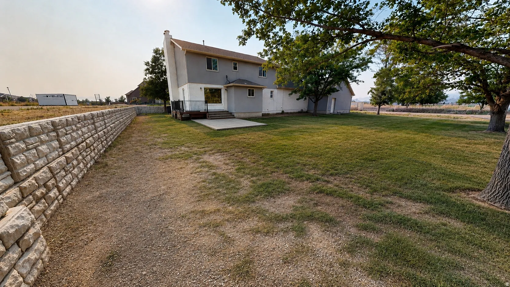 Ai enhanced photo. Expansive backyard featuring a large grass area, mature trees, a concrete patio, and a stone retaining wall