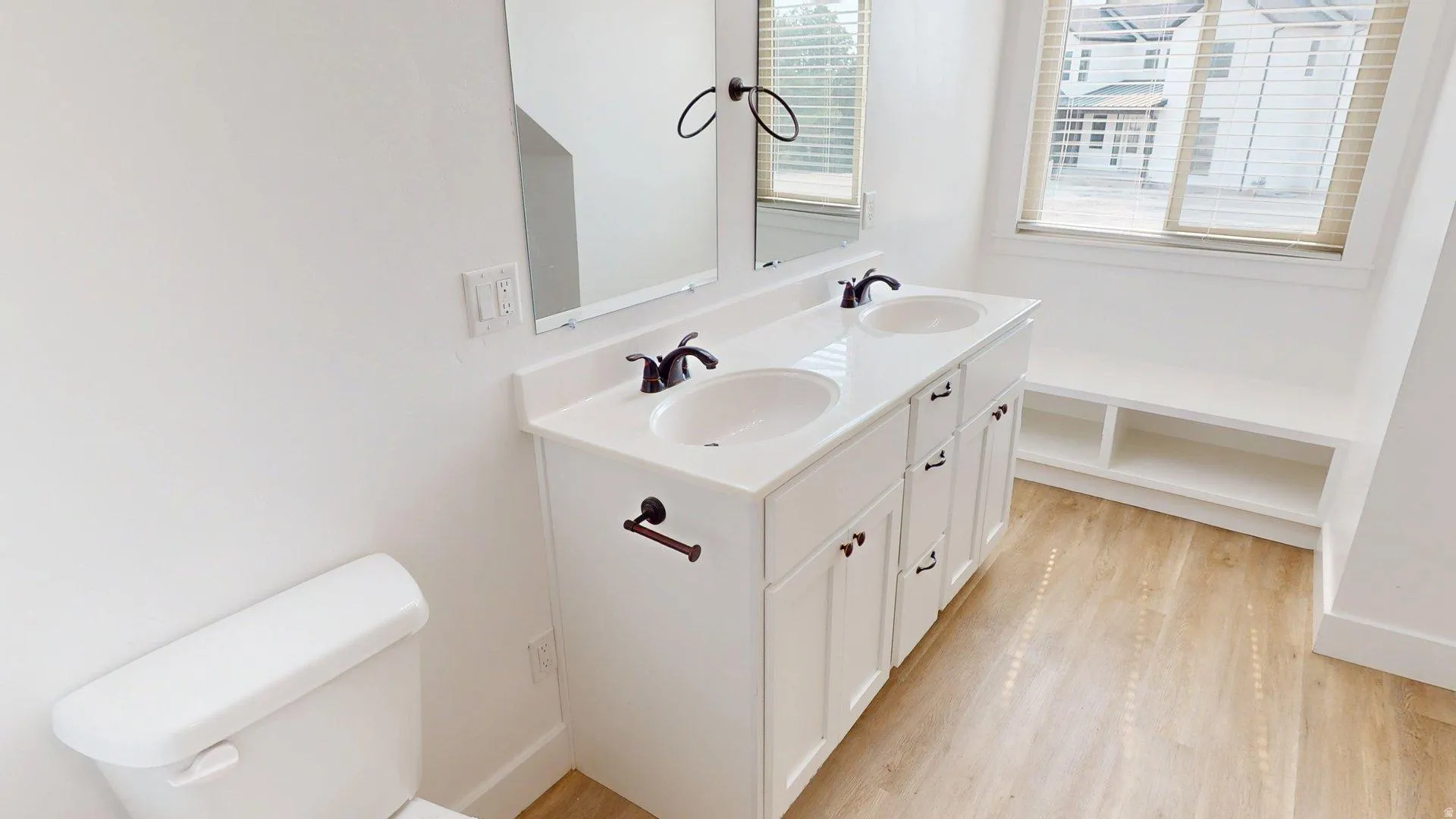 Bathroom featuring a white double vanity with dual sinks and dark bronze fixtures, a built-in window bench with open shelving, wood-finish flooring, and a white toilet