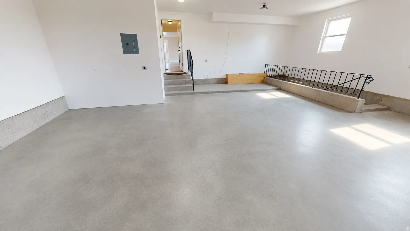 Ai enhanced photo. Spacious concrete-floored garage featuring white walls, an electrical panel, and a stairway with a black metal railing leading to an interior doorway