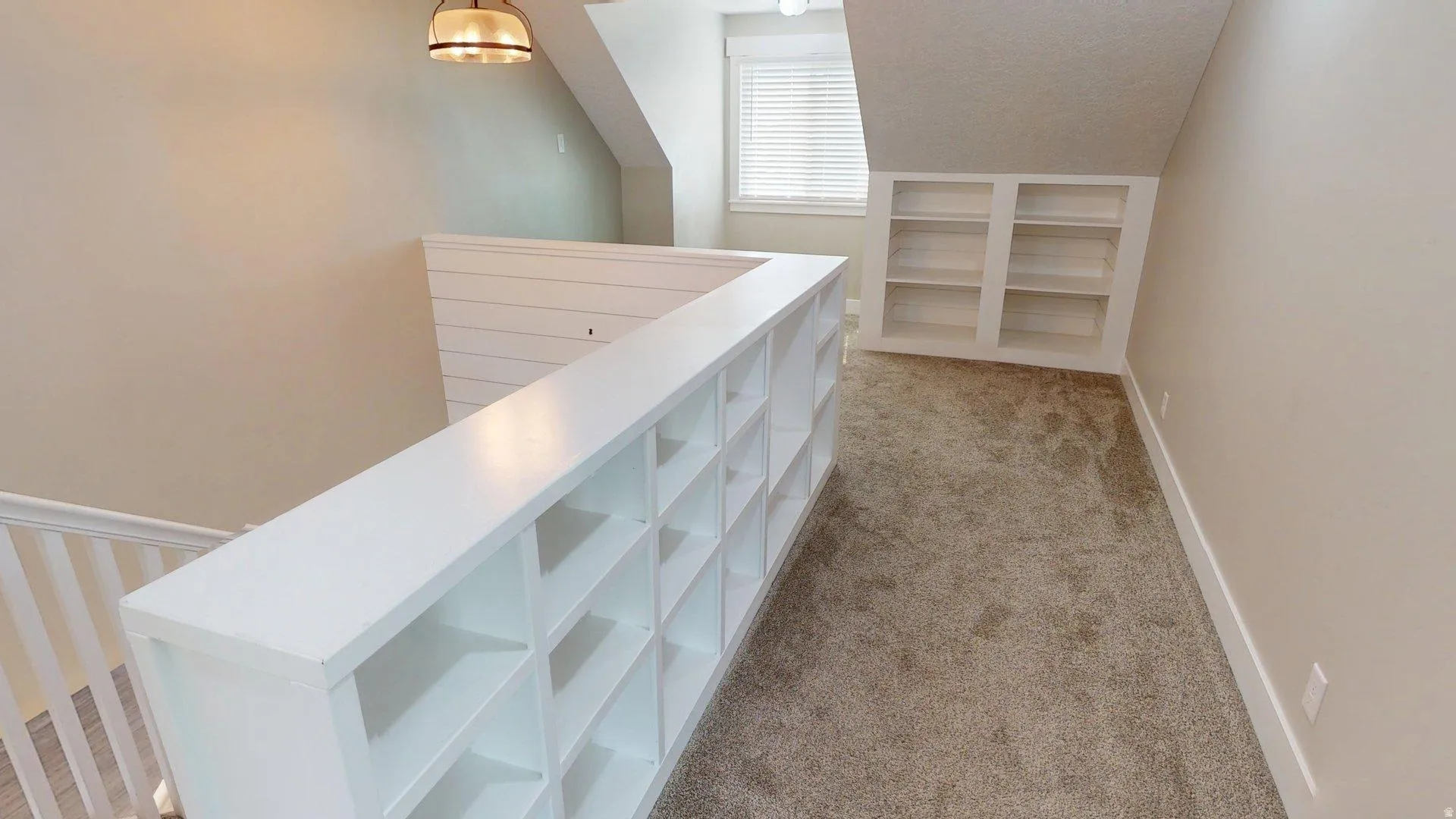 Carpeted upper-level landing featuring built-in white shelving units, a white railing, and a window with blinds