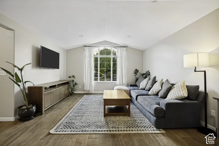 Spacious living area featuring wood-finish flooring, vaulted ceilings, and a large window with white drapery