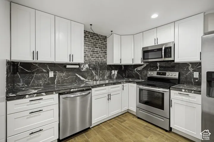 Contemporary kitchen featuring white cabinetry with dark hardware, dark marbled countertops and backsplash, stainless steel appliances, and wood-finish flooring