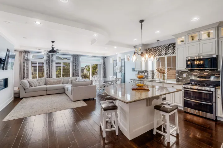 Open-concept living space featuring wood-finish flooring, a contemporary kitchen island with a stone countertop, white cabinetry with integrated lighting, stainless steel appliances, and a recessed fireplace
