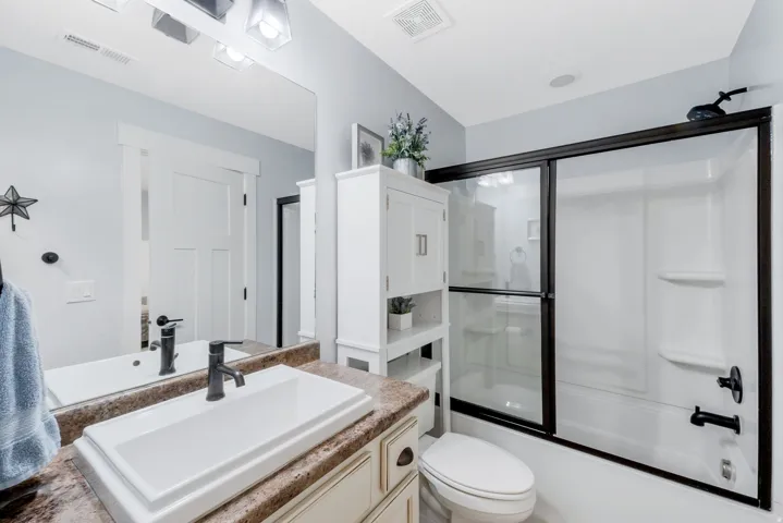 Bathroom featuring a white rectangular vessel sink with a dark bronze-finish faucet, a built-in vanity with a stone-finish countertop, a white freestanding storage cabinet, and a shower-tub combination with dark bronze-finish sliding glass doors