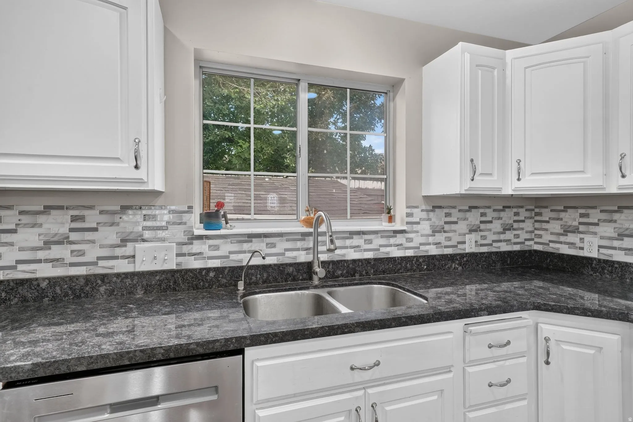 Kitchen featuring white cabinetry, dark speckled countertops, a tile backsplash, stainless steel double-basin sink, and a window with grid detailing
