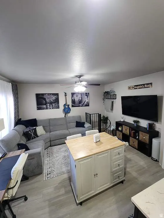 Open-concept living space featuring light wood-finish flooring, a ceiling fan with integrated lighting, a contrasting black spiral staircase, and a light-toned kitchen island with a wood-finish countertop