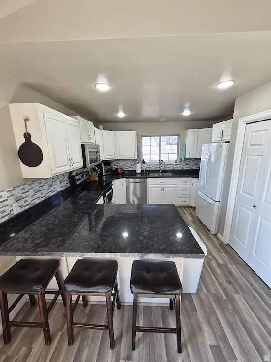 Bright kitchen featuring white cabinetry, dark granite countertops, stainless steel appliances, a tiled backsplash, and wood-finish flooring