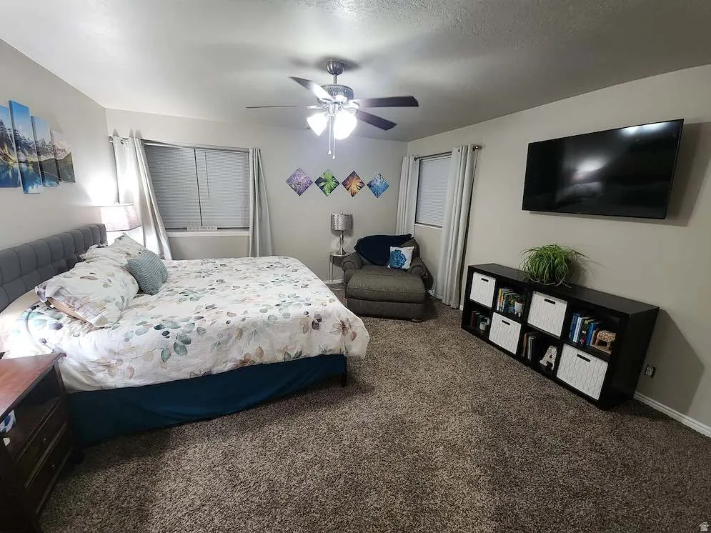 Carpeted bedroom featuring multiple windows, a ceiling fan with integrated lighting, and a wall-mounted display