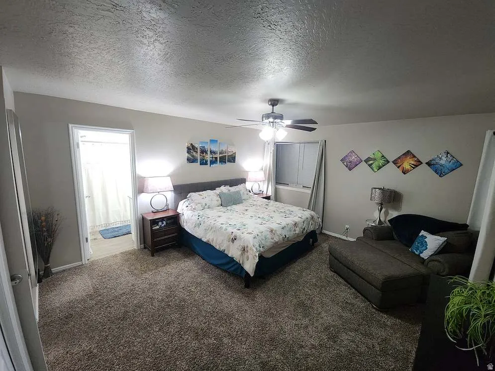 Carpeted room featuring a ceiling fan, window with vertical blinds, and an open doorway revealing a bathroom with wood-finish flooring and a shower curtain