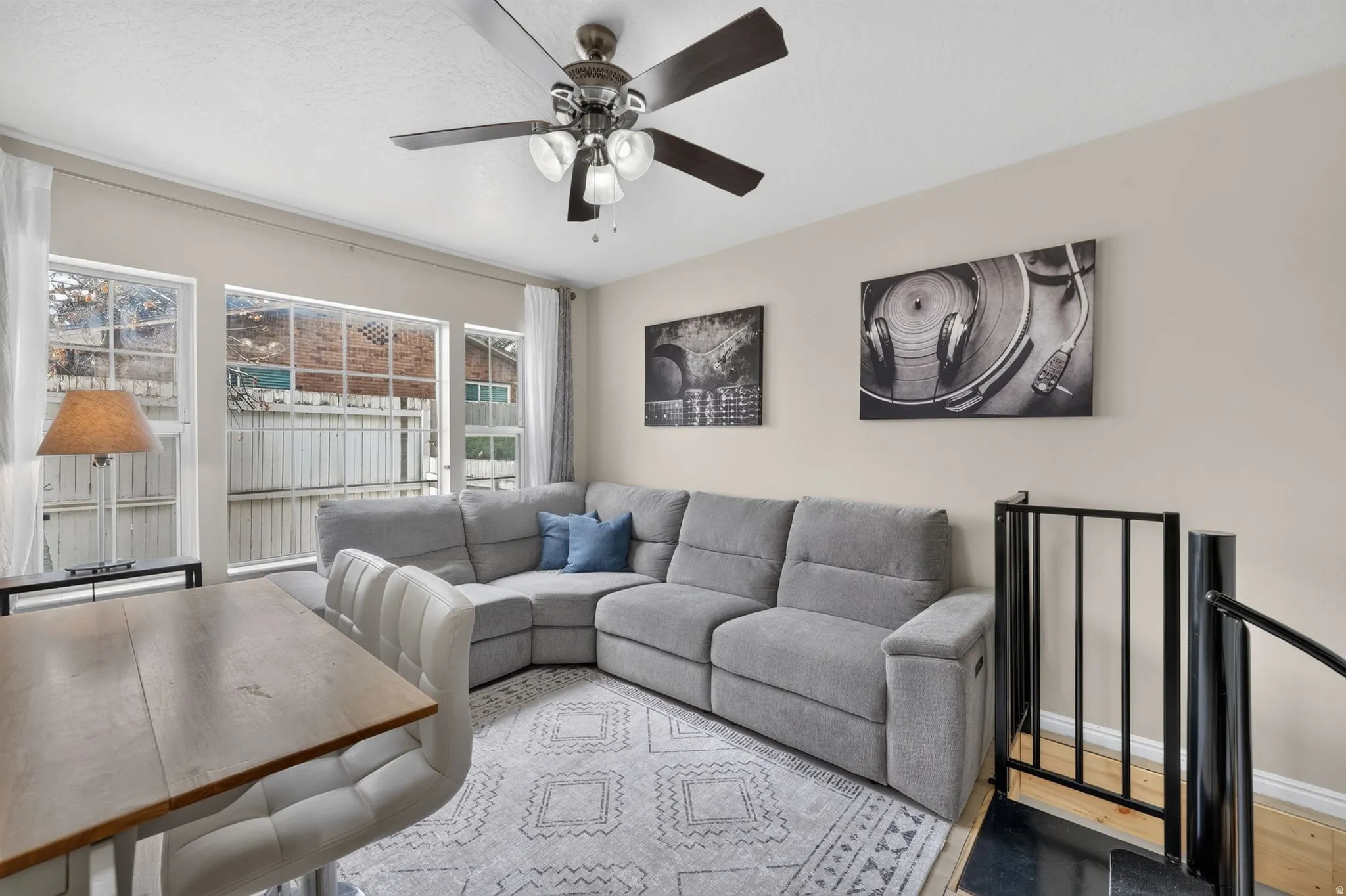 Living area featuring multiple windows, a ceiling fan with integrated lighting, and a partial view of a staircase with black metal railings