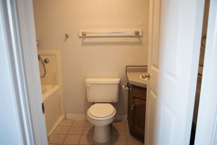 Bathroom featuring a white toilet, an integrated bathtub and shower with a flexible shower head, a wood-finish vanity with a light-toned countertop, and light-toned tile flooring